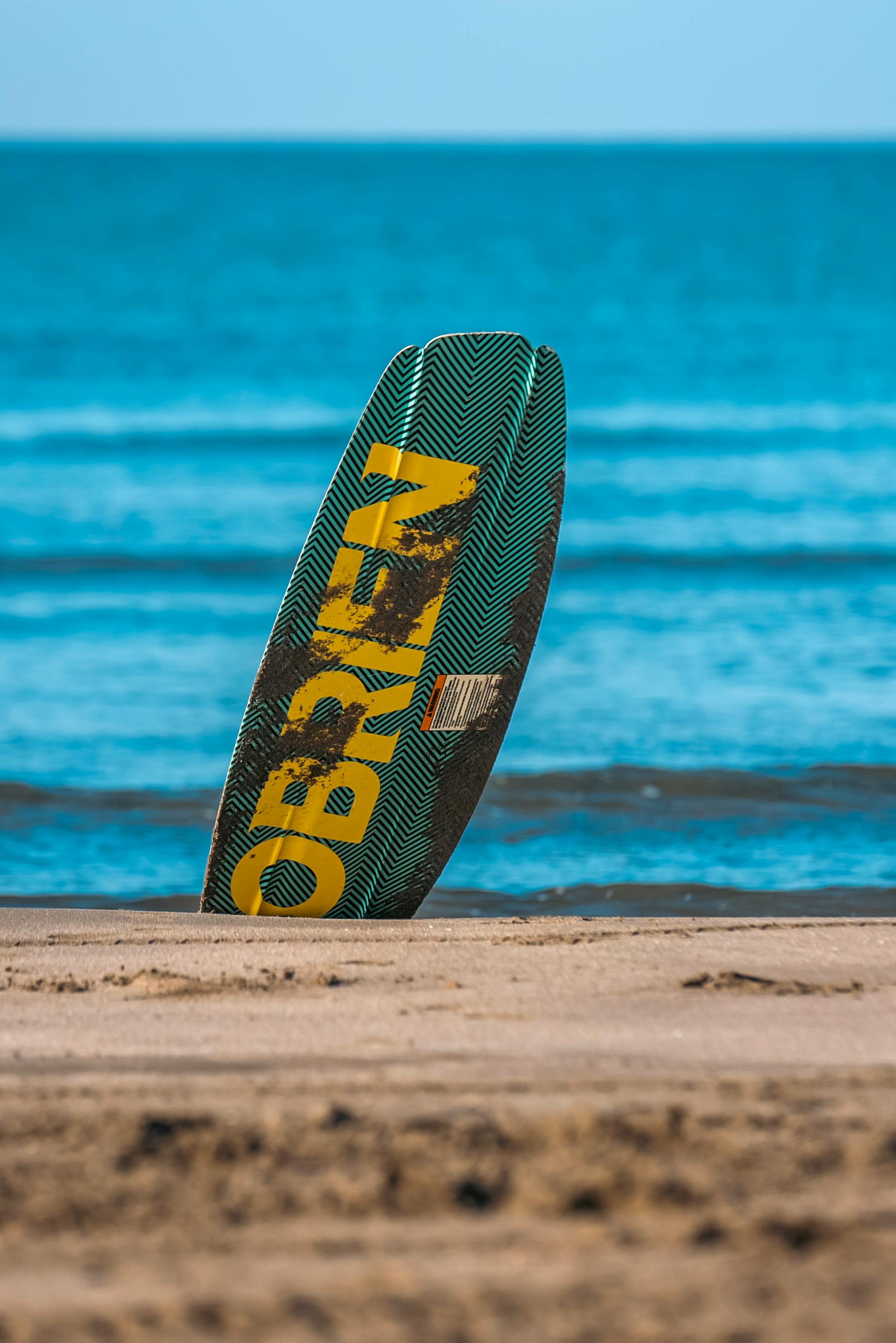 A surfboard stands upright on the sandy beach of Antalya, Türkiye, against a backdrop of the turquoise sea.