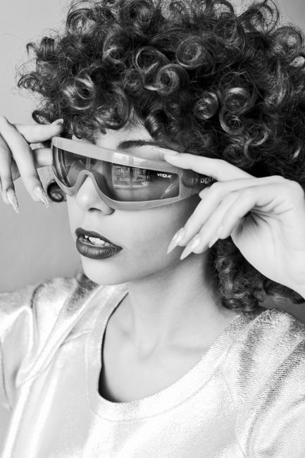 Black and white portrait of a woman with curly hair adjusting her sunglasses, exuding chic style.