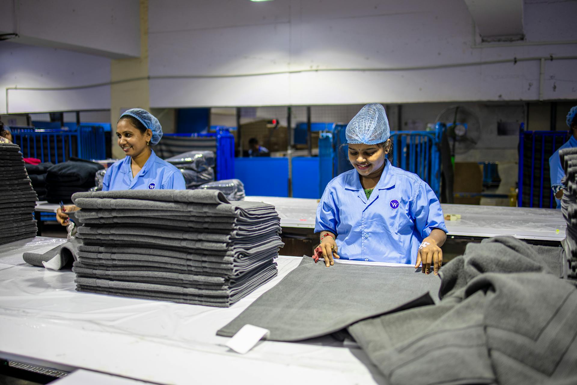 Two female textile workers folding fabrics in an indoor factory setting, wearing blue uniforms and hair nets.