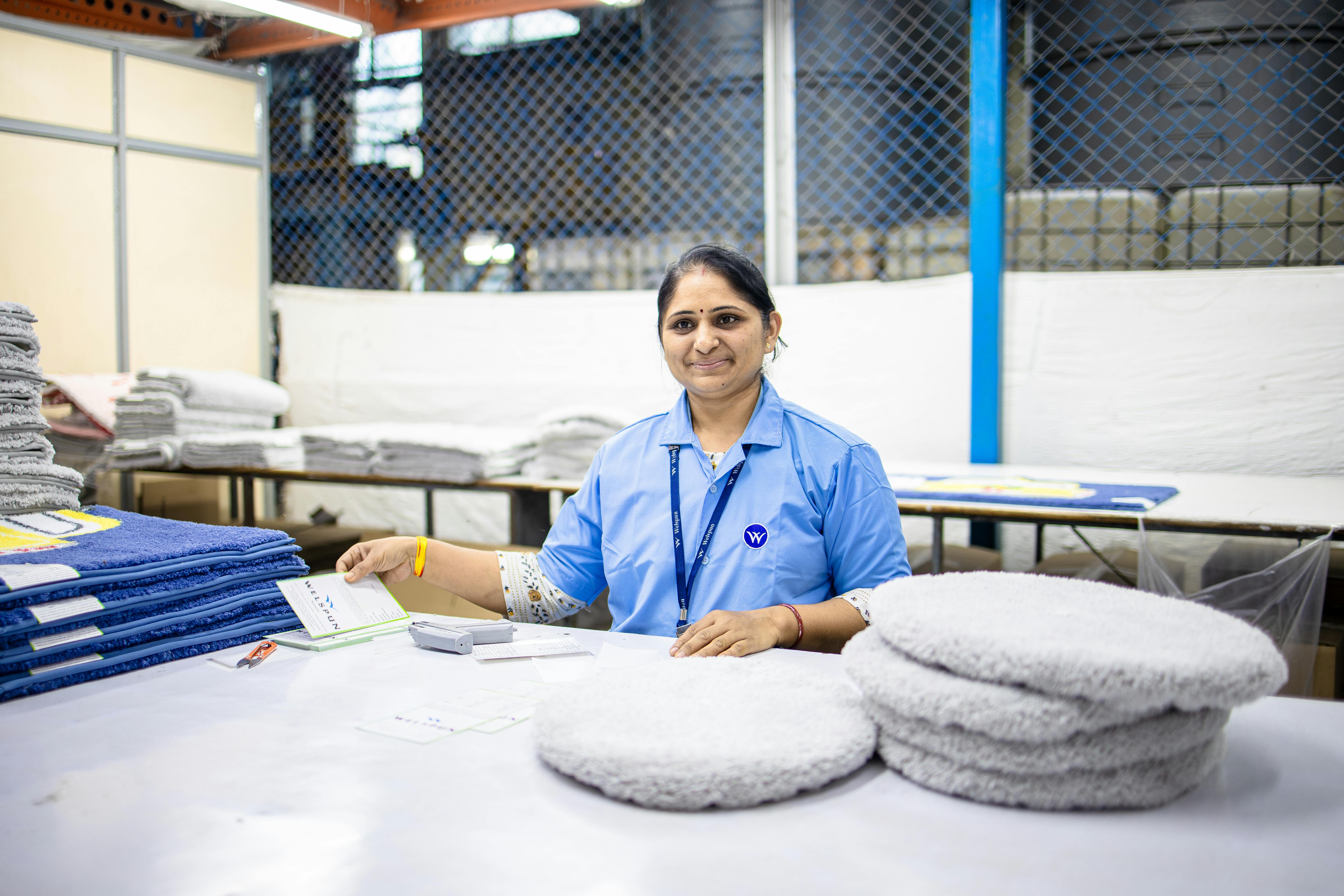 Female textile worker smiling in a factory surrounded by fabrics and packaged products.