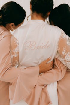 Bride embraces bridesmaids in elegant lace robes, wedding preparation moment.