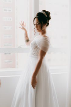 Elegant bride in white dress and tiara posing by a sunlit window indoors.