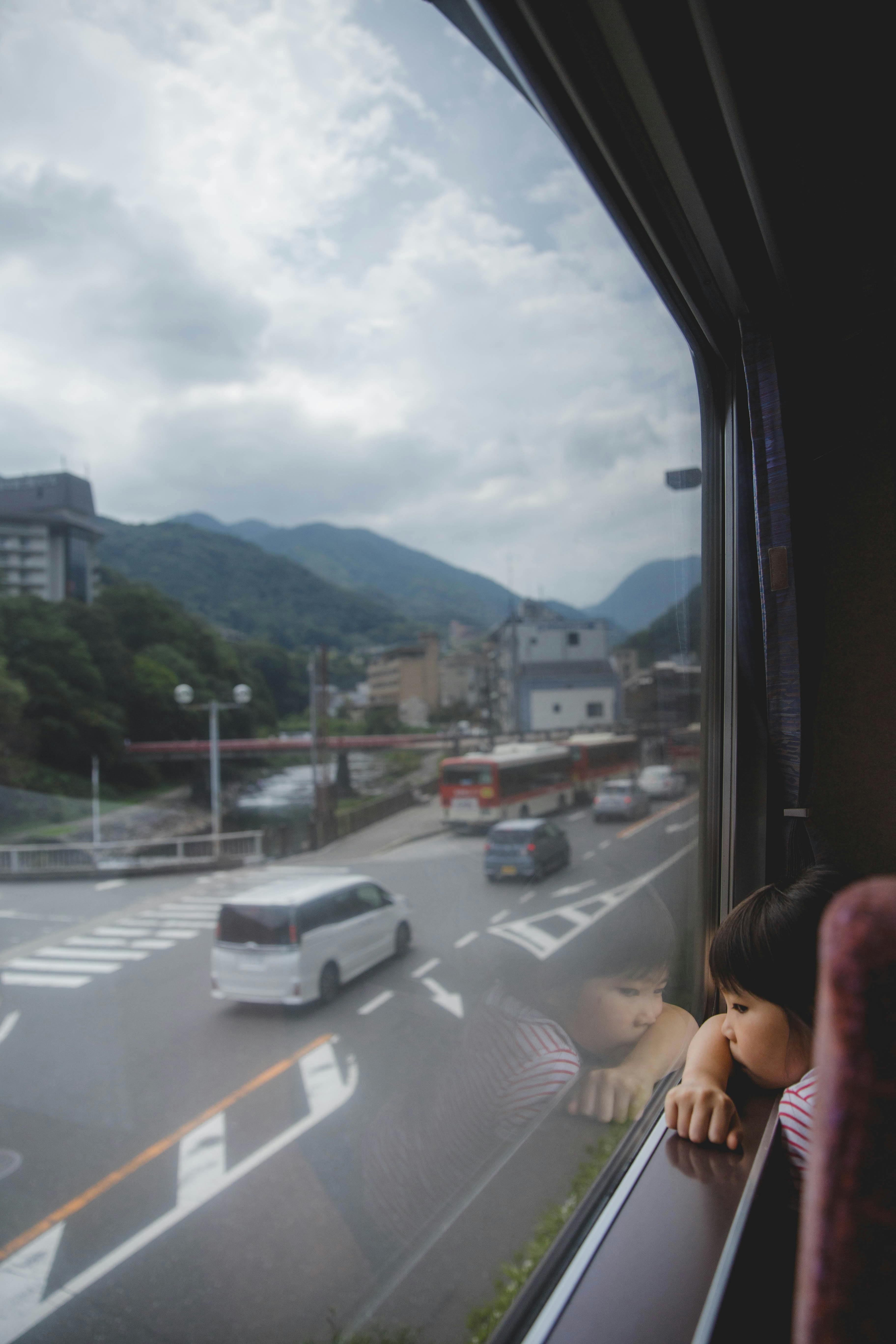 A child gazes out a train window at a Japanese landscape with mountains and city streets.