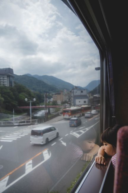 A child gazes out a train window at a Japanese landscape with mountains and city streets.