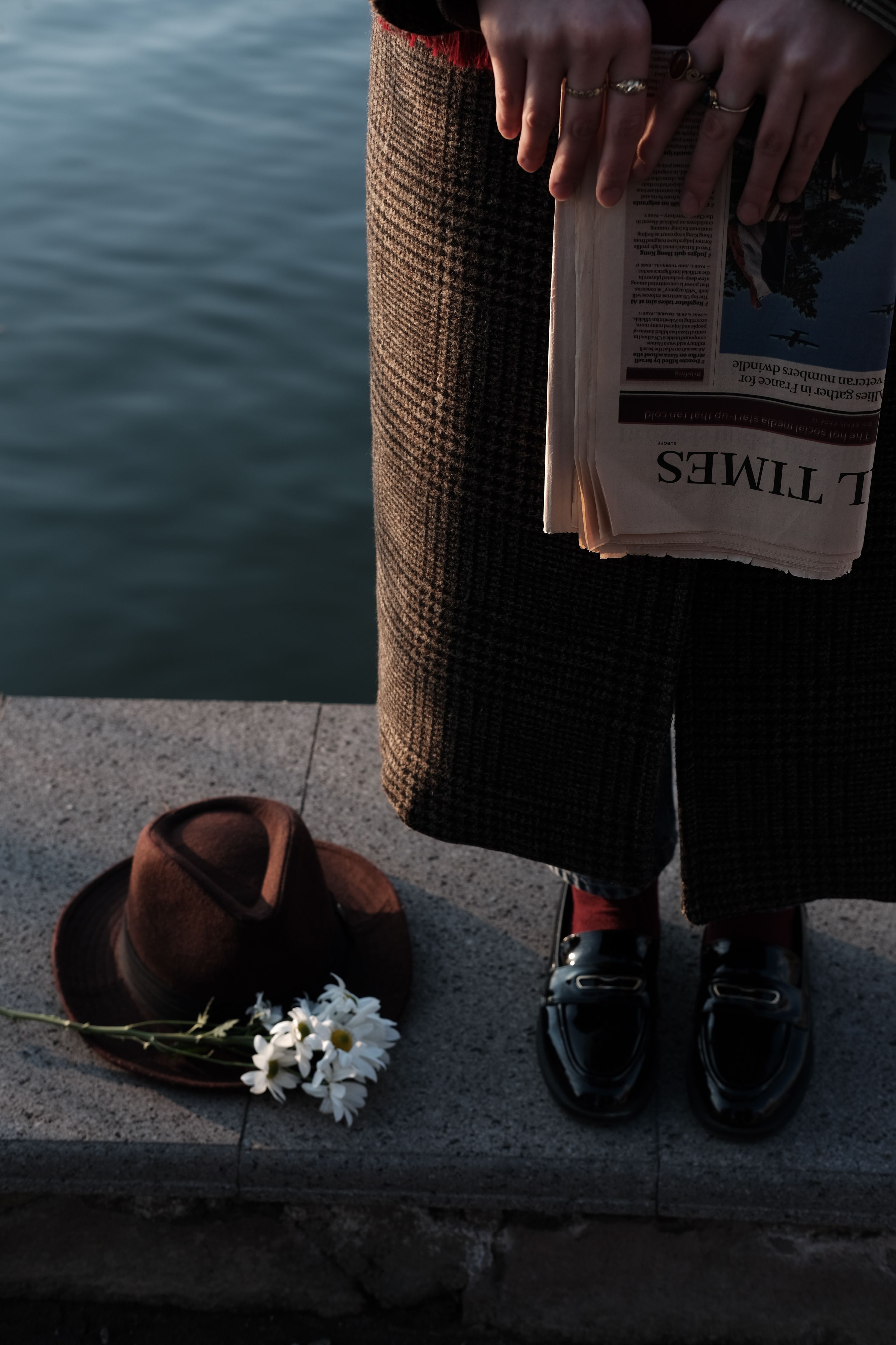 Pessoa segurando um jornal com flores e um chapéu no chão ao lado da água.