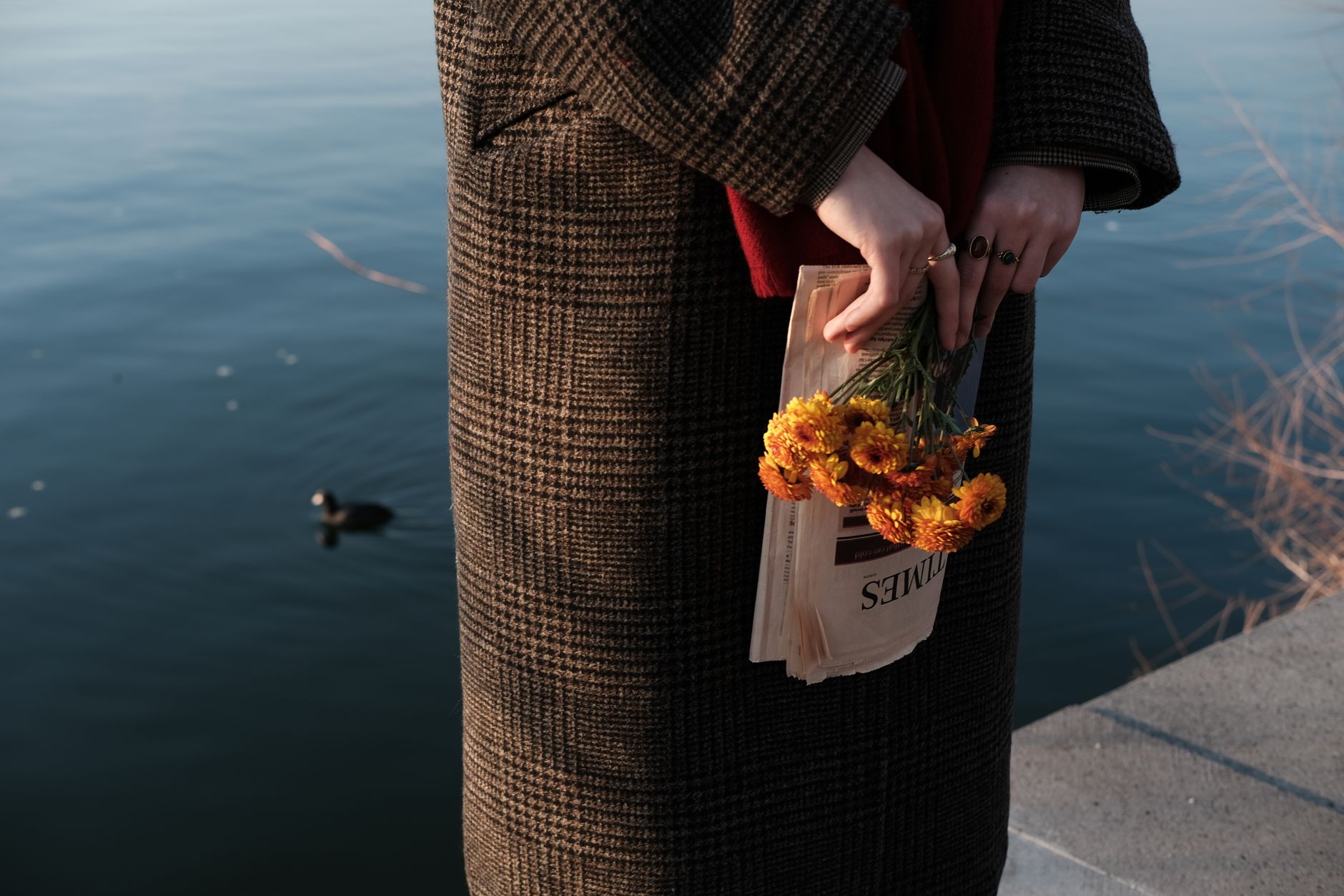 A woman in a coat holds yellow flowers and a newspaper by a calm lake, capturing a serene moment.