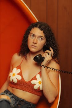 Woman with curly hair in a daisy crochet top using a vintage phone in a retro setting.