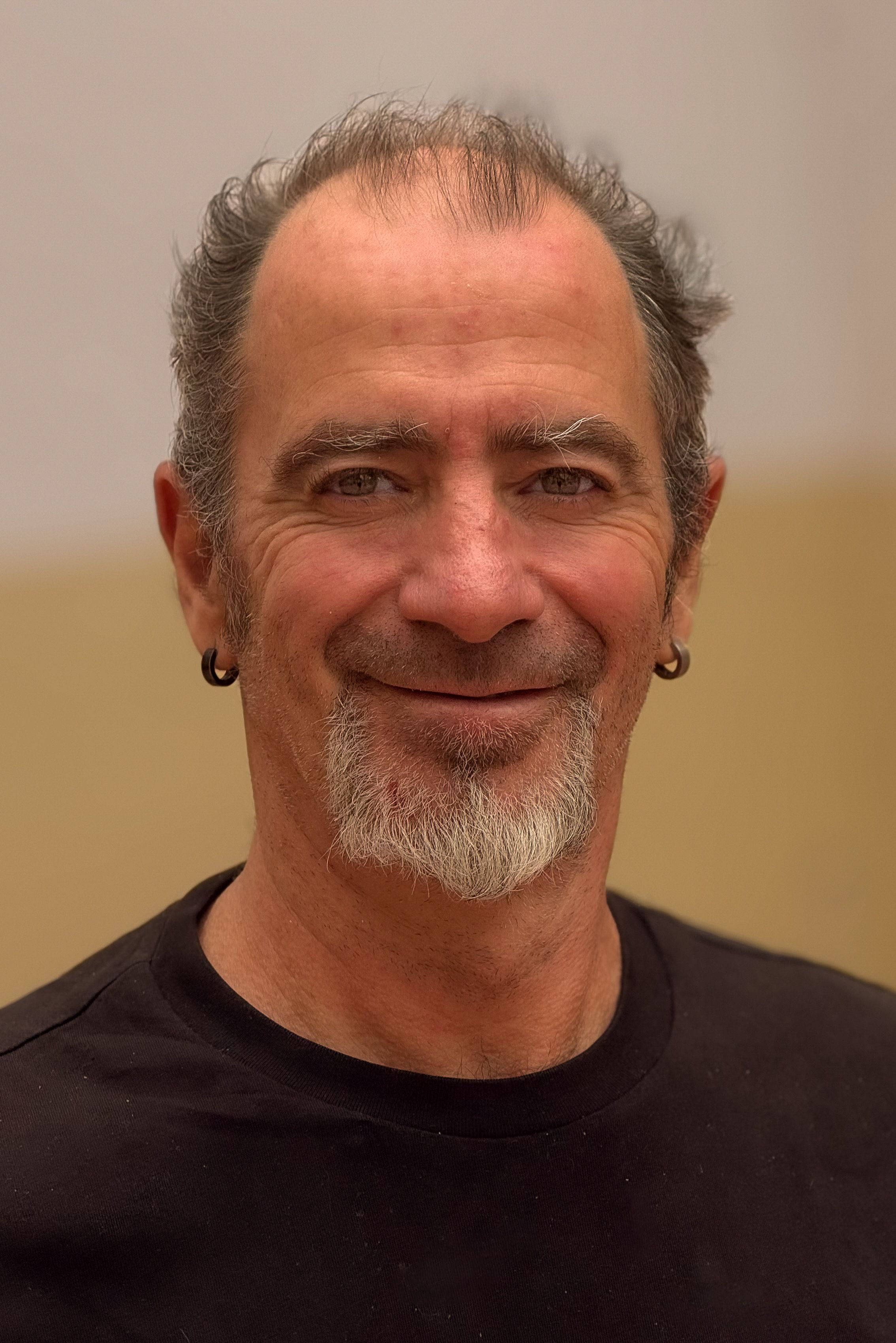 Close-up portrait of a smiling mature man with earrings and facial hair, wearing a black shirt.