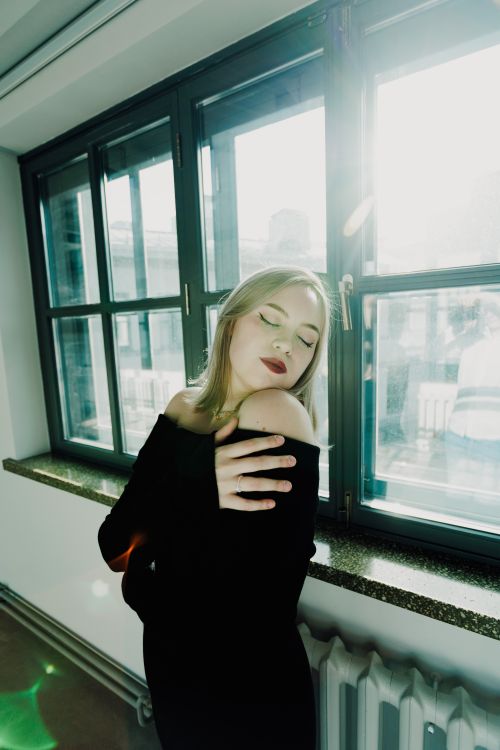 Free Elegant woman in black dress with red lipstick posing by a sunlit window indoors. Stock Photo