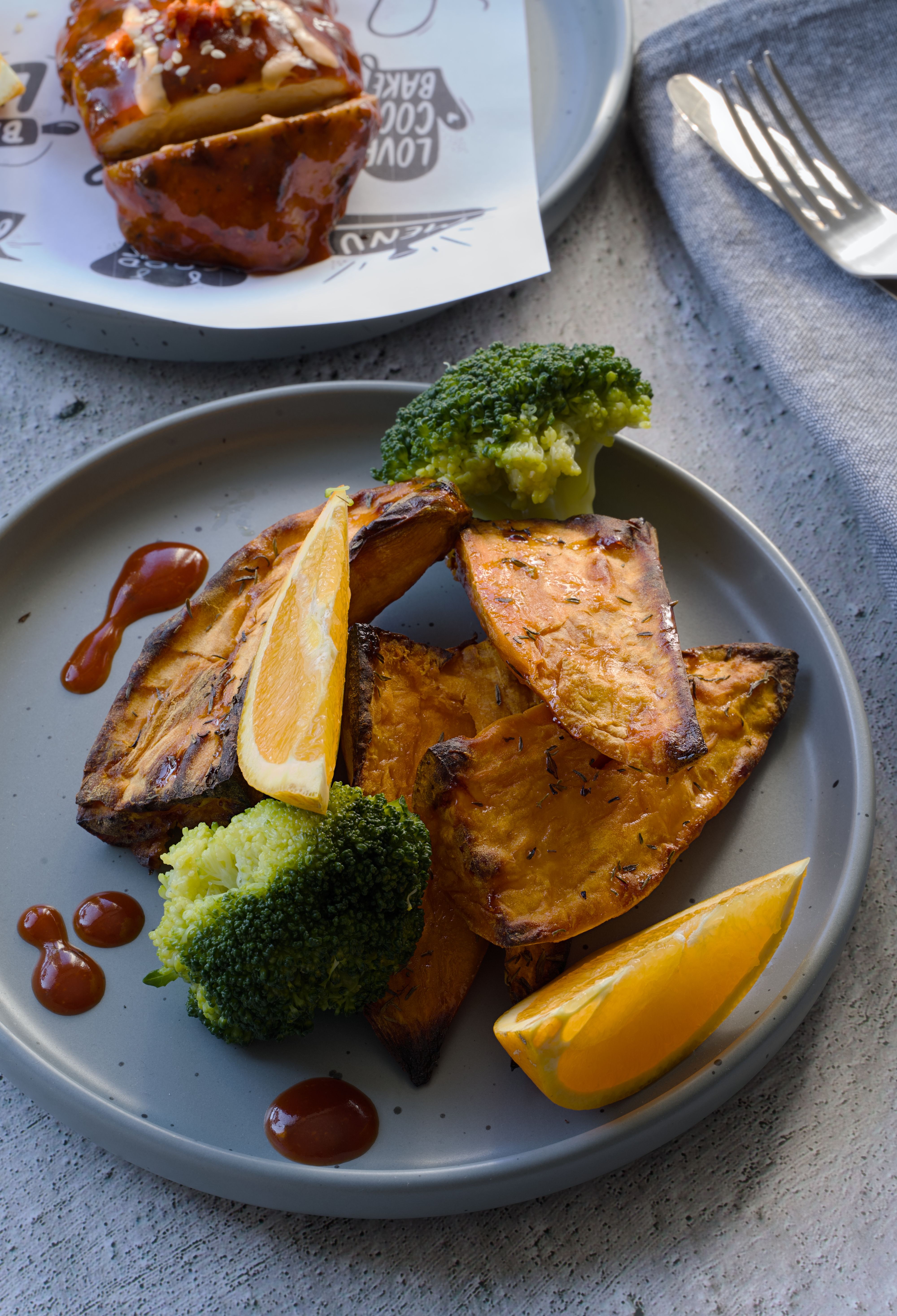 Four glass meal prep containers filled with roasted chicken thighs, cubed sweet potato, charred broccoli florets, and sliced avocado, with a small jar of lemon dressing beside them.