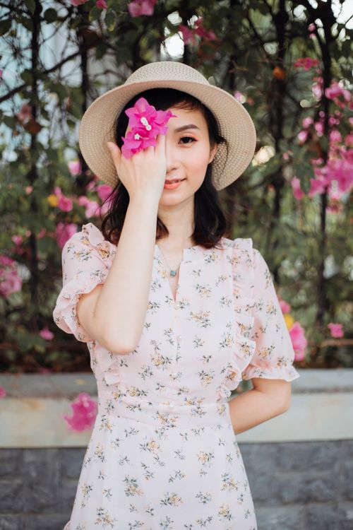 Free A woman in a floral dress and sun hat playfully holds a pink flower, smiling outdoors. Stock Photo