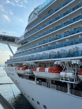 A luxury cruise ship with lifeboats docked at an ocean port under a clear sky.
