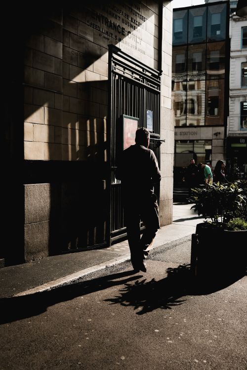 Free A man walks in the shade towards a sunlit city street, casting a shadow. Stock Photo