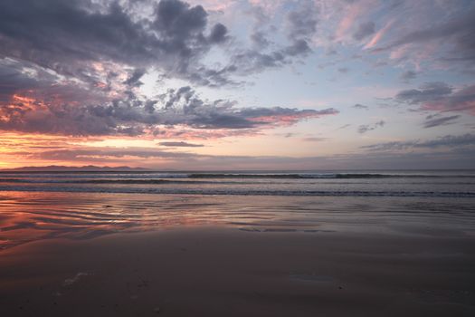 A tranquil sunset over the ocean with vibrant clouds and reflective waves on the sand.