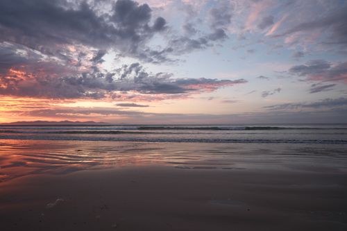 Free A tranquil sunset over the ocean with vibrant clouds and reflective waves on the sand. Stock Photo