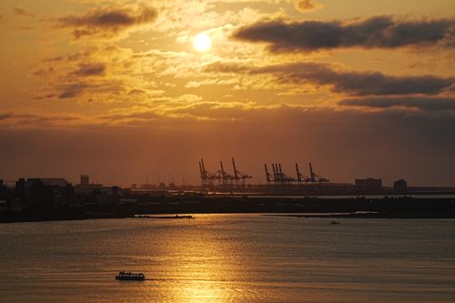 Free Sunset scene with cranes silhouetted against the sky over a harbor, casting reflections on the water. Stock Photo