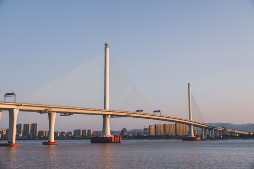 Free Suspension bridge spanning a river at sunset, with city skyline and mountains in the background. Stock Photo