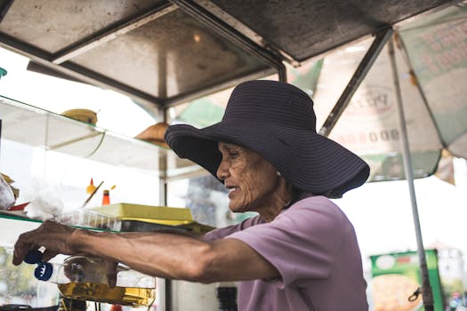 An elderly woman wearing a wide-brimmed hat selling goods at an outdoor market stall.