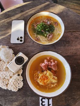 Two noodle bowls with fresh toppings and broth on a rustic table with a vintage camera.