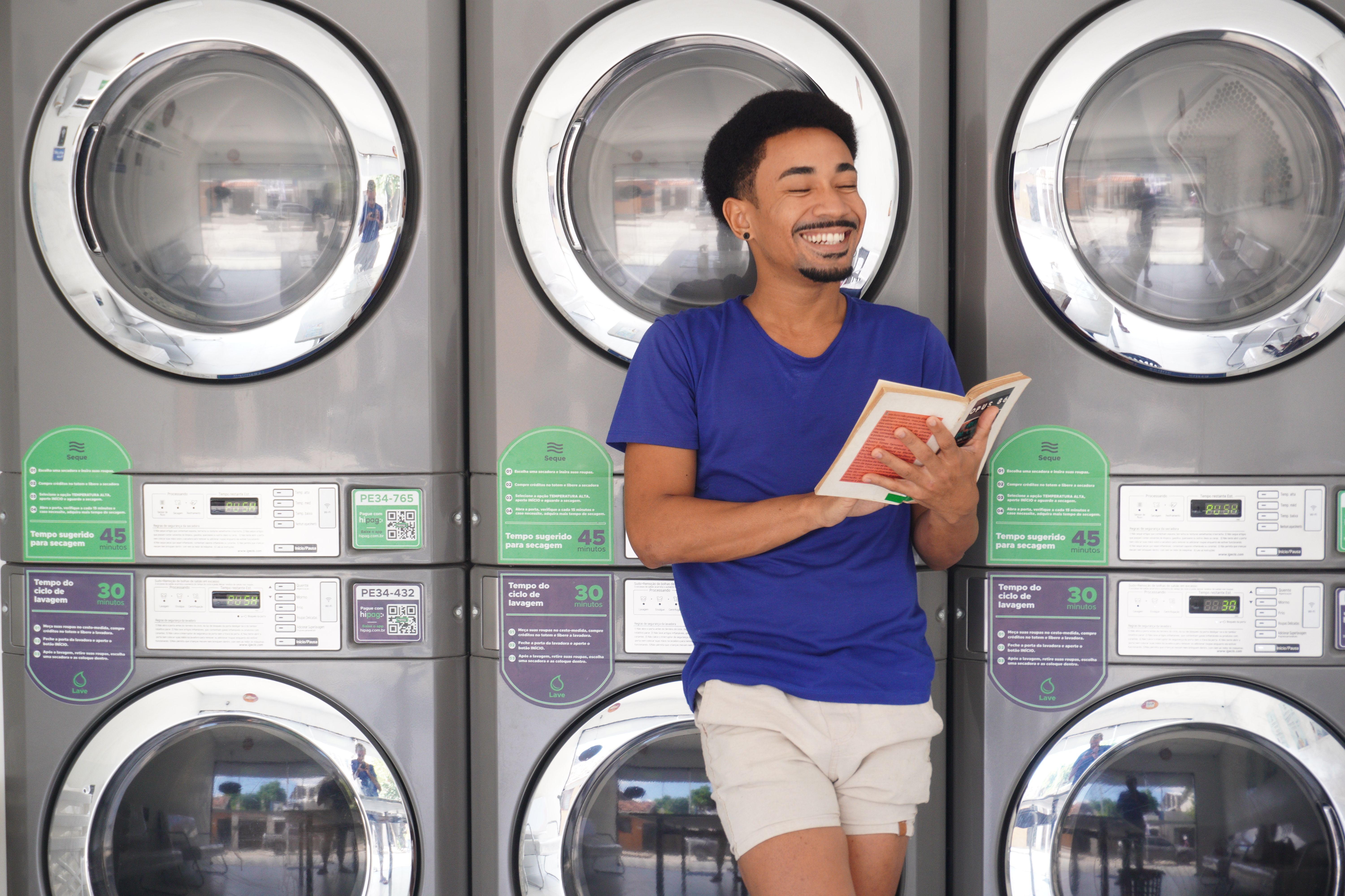 Homem sorridente aprecia um livro enquanto lava roupa em uma lavanderia brasileira.