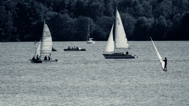 A serene view of sailboats and a windsurfer on a calm lake in Saarland, Germany, surrounded by lush forests.