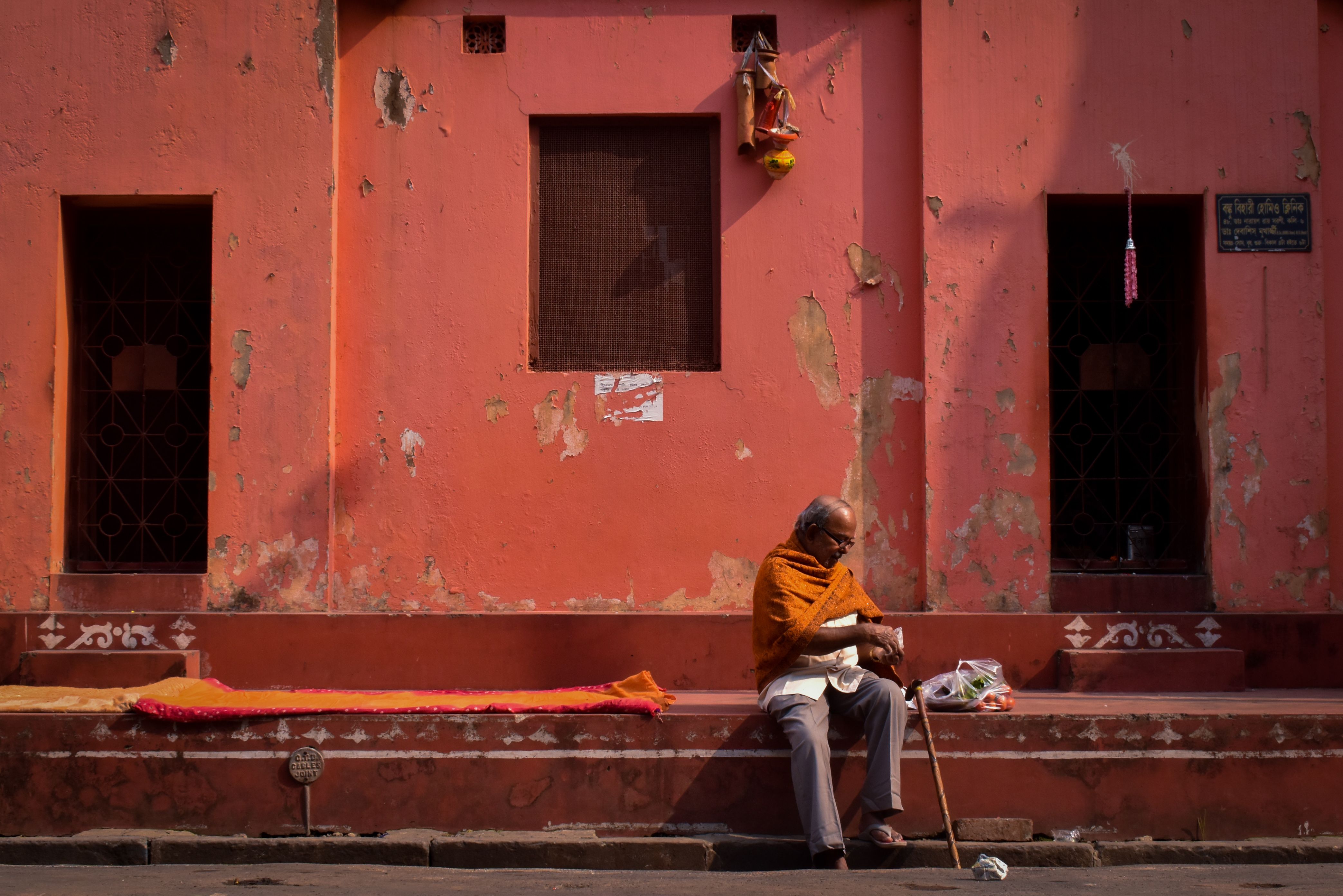 A senior man sits wrapped in an orange shawl against a rustic wall in Kolkata, India.