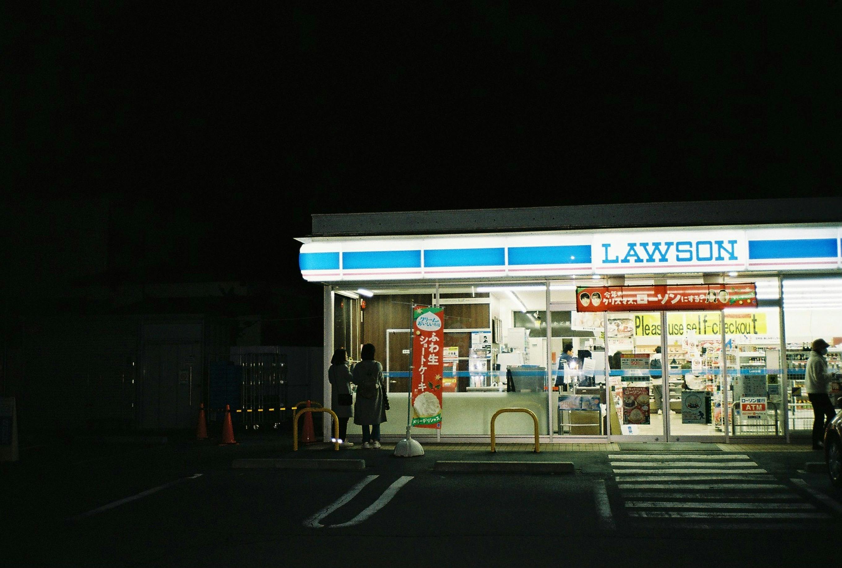 A nighttime view of a Lawson convenience store entrance with people.