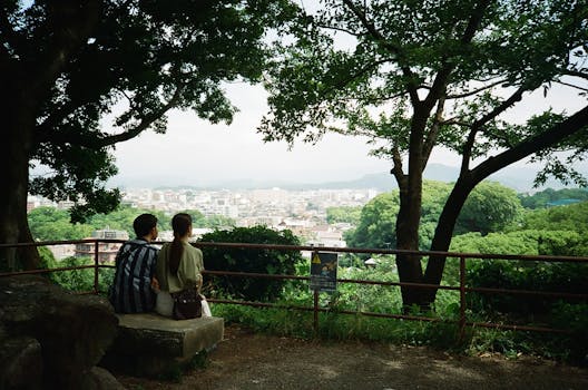 Young couple sitting in a park, overlooking a scenic city view surrounded by lush greenery.