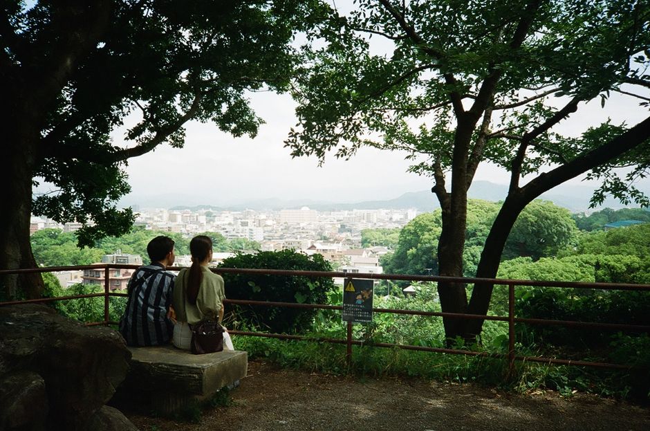 Young couple sitting in a park, overlooking a scenic city view surrounded by lush greenery.