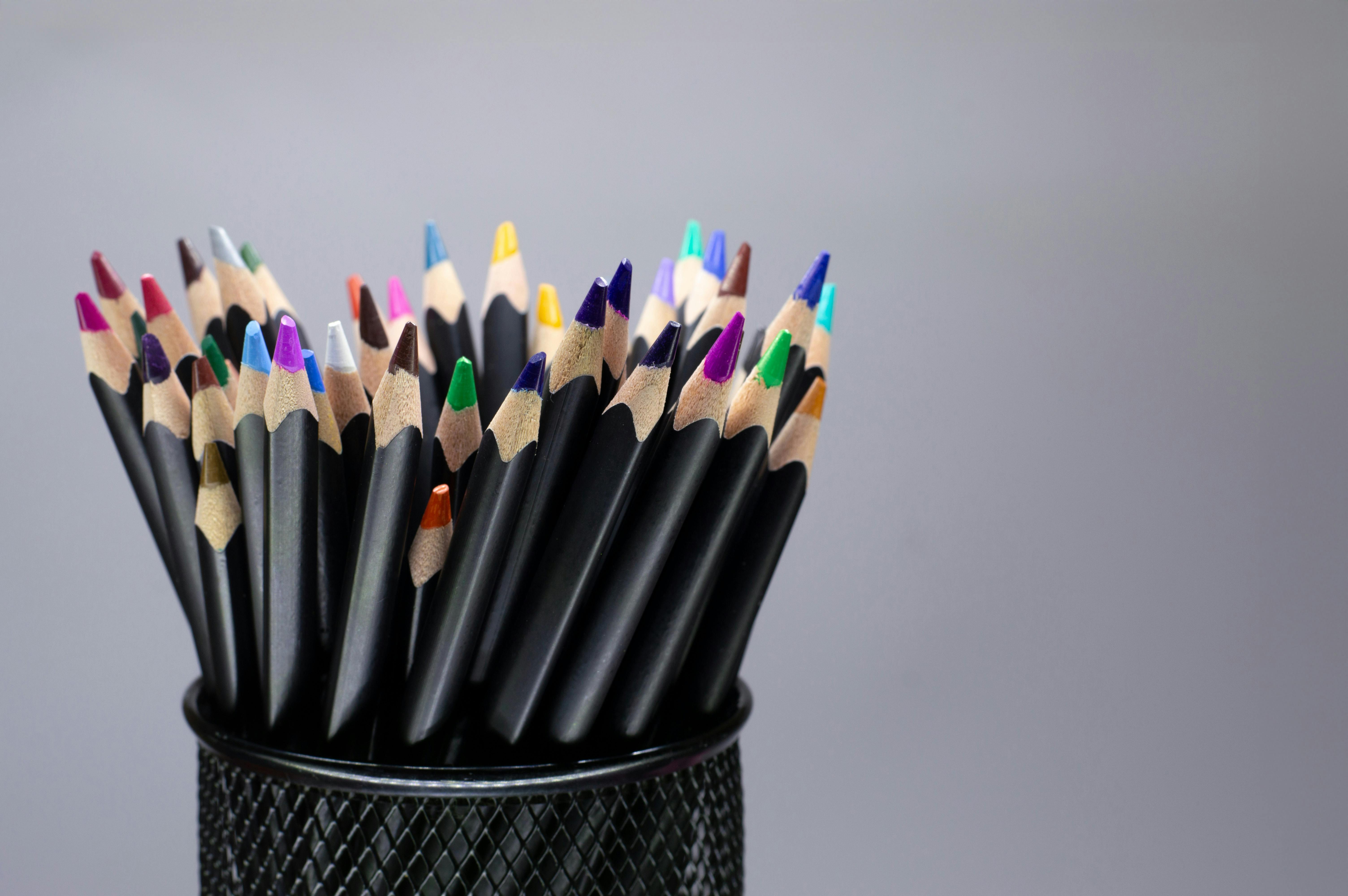 A collection of colorful pencils in a black mesh holder against a neutral background.