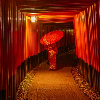 A woman in traditional attire walking with an umbrella under vibrant torii gates at night.（イメージ）