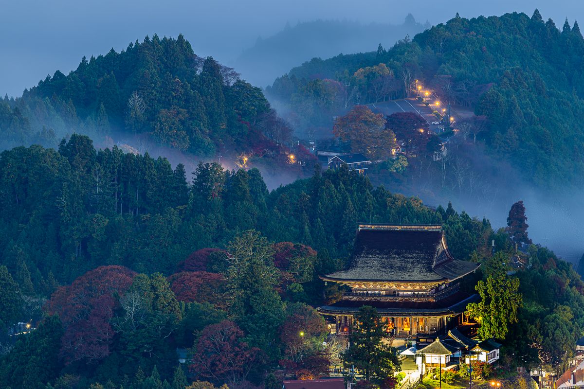 A traditional Japanese temple hall surrounded by mist-covered forested mountains