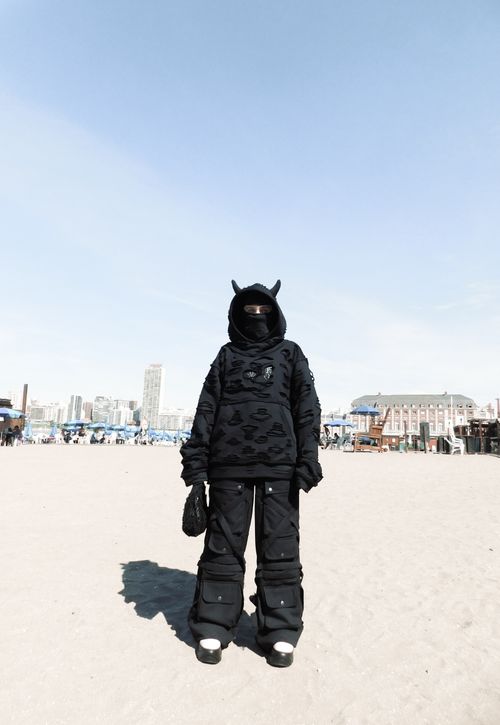 Free An individual dressed in a unique black outfit stands on a beach with a city skyline in the background. Stock Photo