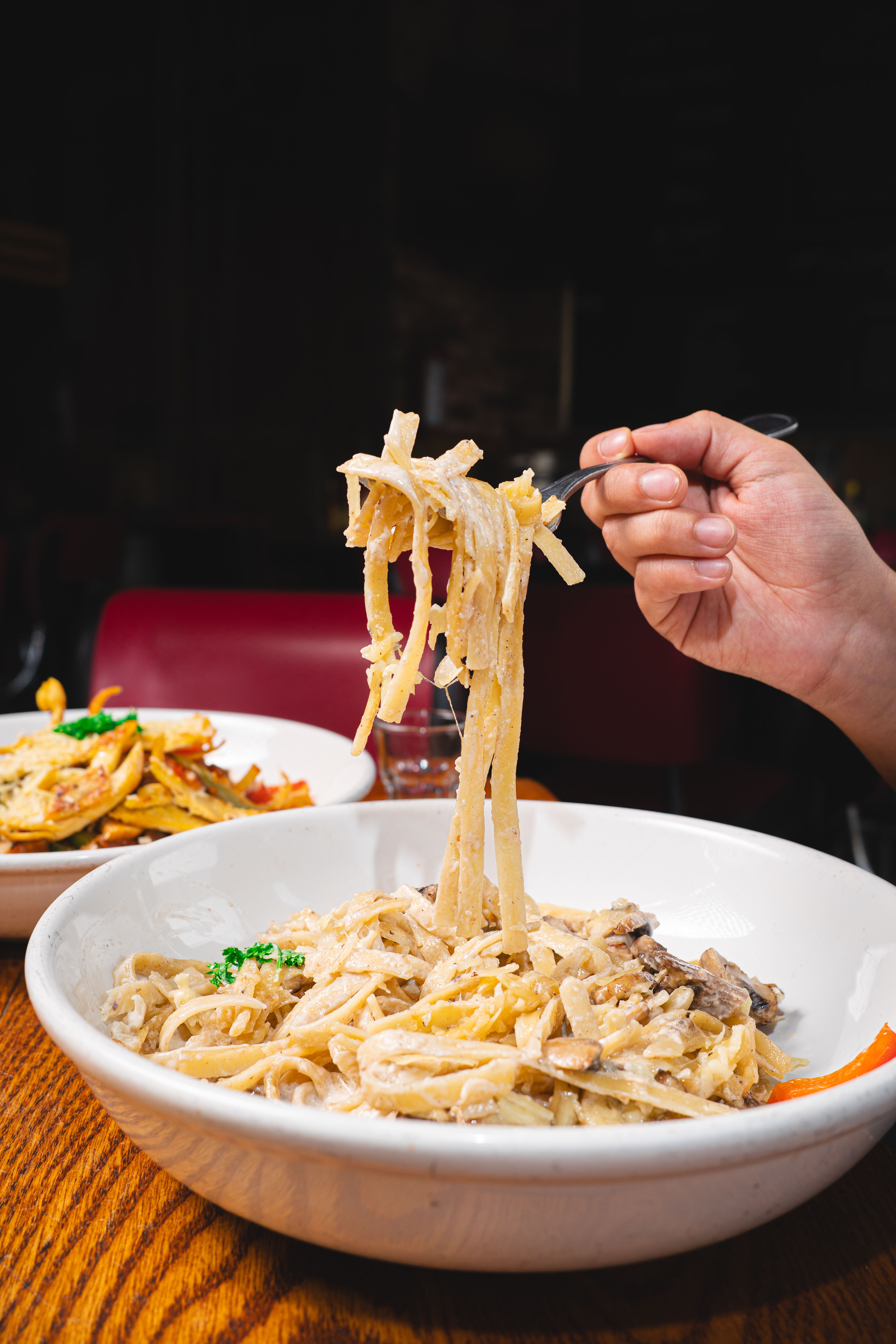 A hand holding a fork with creamy fettuccine Alfredo, garnished with herbs, in a cozy restaurant setting.