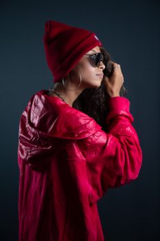 Fashionable woman in red jacket, beanie, and sunglasses against a dark backdrop.