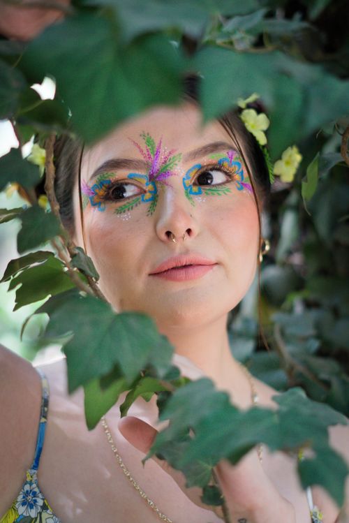 Free A woman with colorful eye makeup peeks through lush green leaves outdoors. Stock Photo
