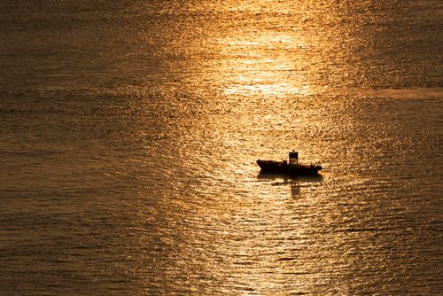 Free A lone boat silhouetted against the shimmering gold sea during sunset, creating a serene and picturesque scene. Stock Photo