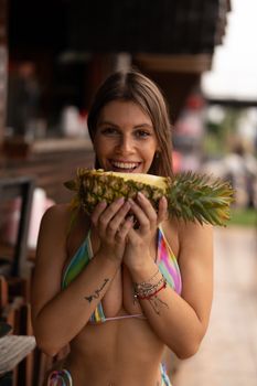 Woman in colorful bikini smiling and holding a pineapple outdoors, embodying tropical vibes and joy.