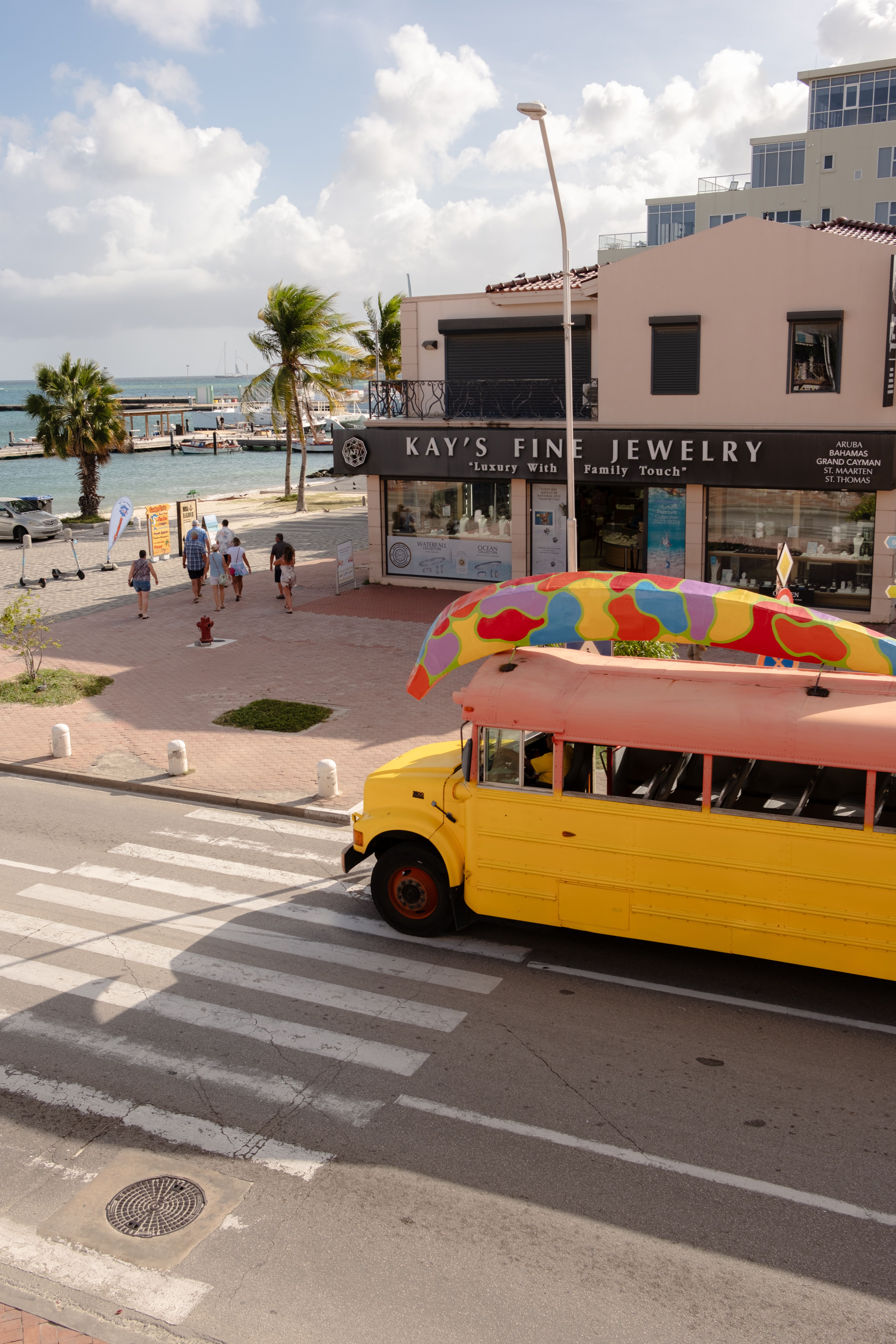 A public passenger bus moving through a sunny Caribbean street, with pedestrians and palm trees
