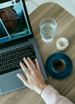 Overhead view of a hand on a laptop next to coffee and water, cozy desk setup.