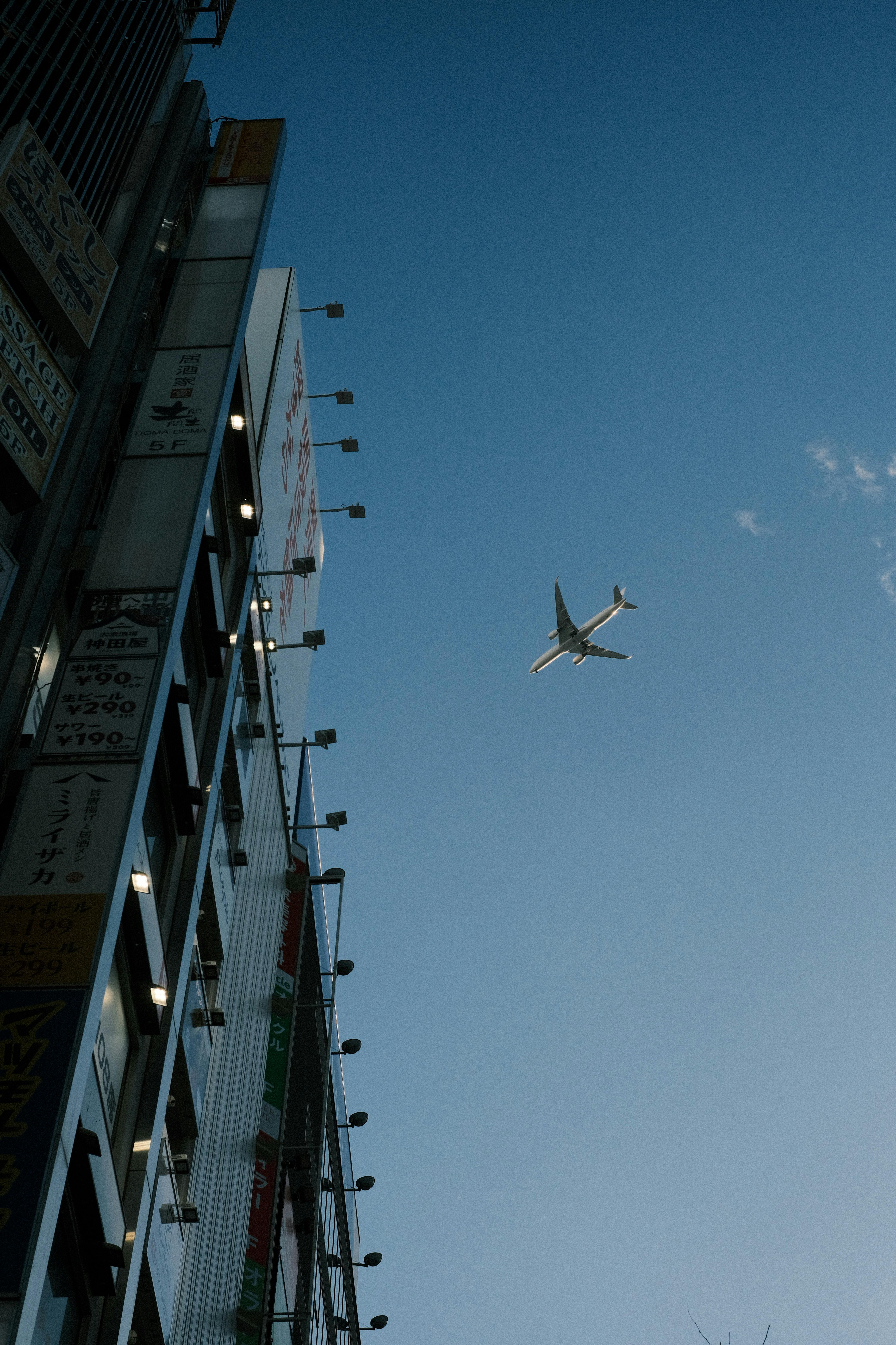 A commercial airplane flies over a tall building in Tokyo, Japan, silhouetted against a bright blue sky.