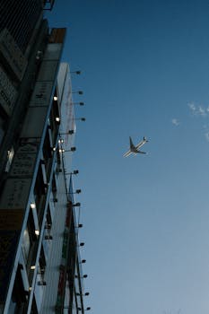 A commercial airplane flies over a tall building in Tokyo, Japan, silhouetted against a bright blue sky.