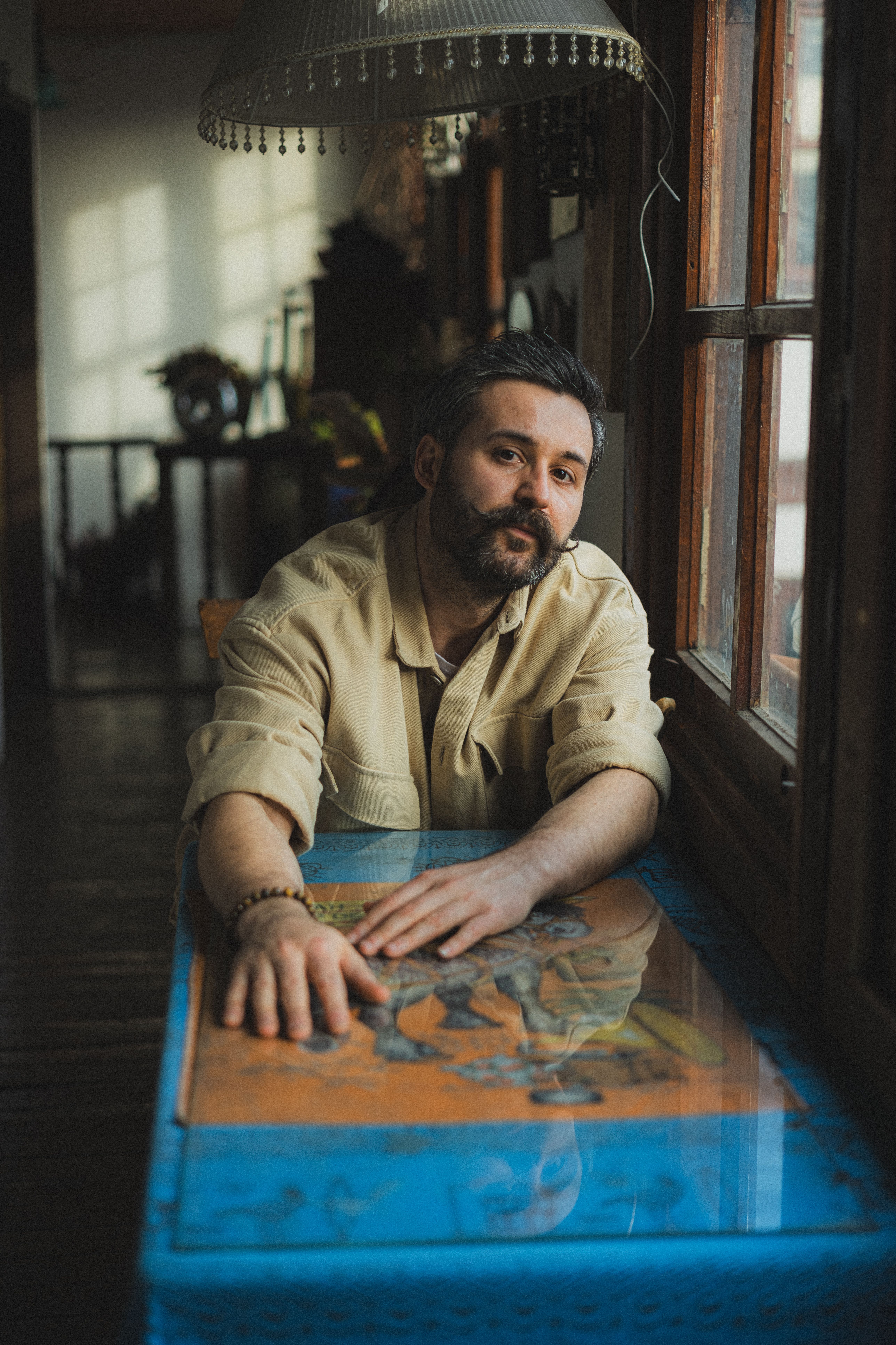 Man in casual attire sitting by a window in a cozy indoor setting, reflecting.