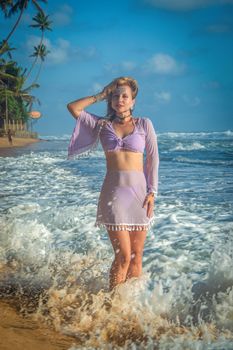 A woman in a lavender outfit standing by the ocean waves on a sunny tropical beach.