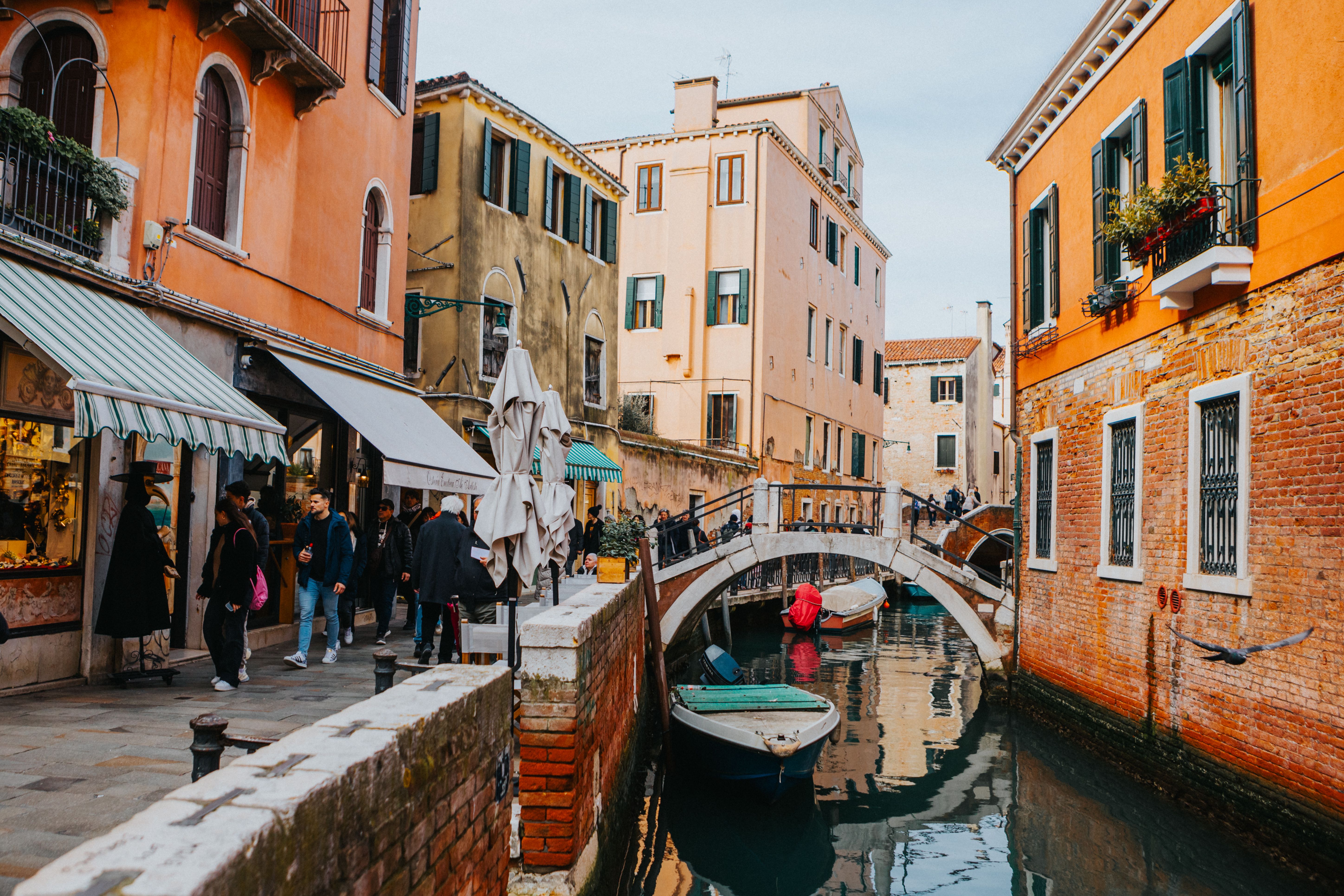 A narrow canal with a small stone bridge, moored boats and orange and ochre brick buildings, with shop awnings and a few pedestrians on the left bank