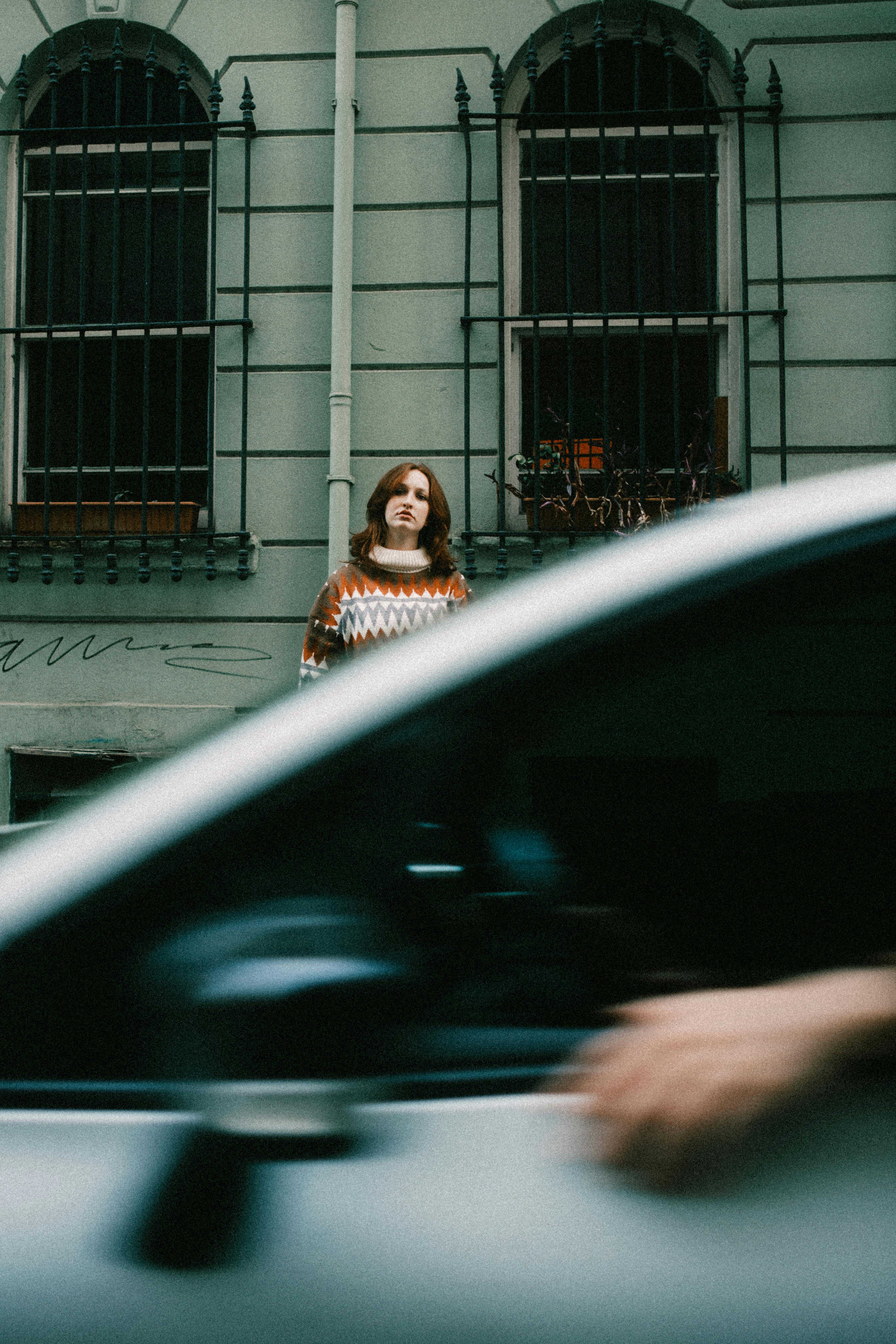 A woman in a colorful sweater stands behind a moving car in an urban street, evoking a sense of motion and city life.