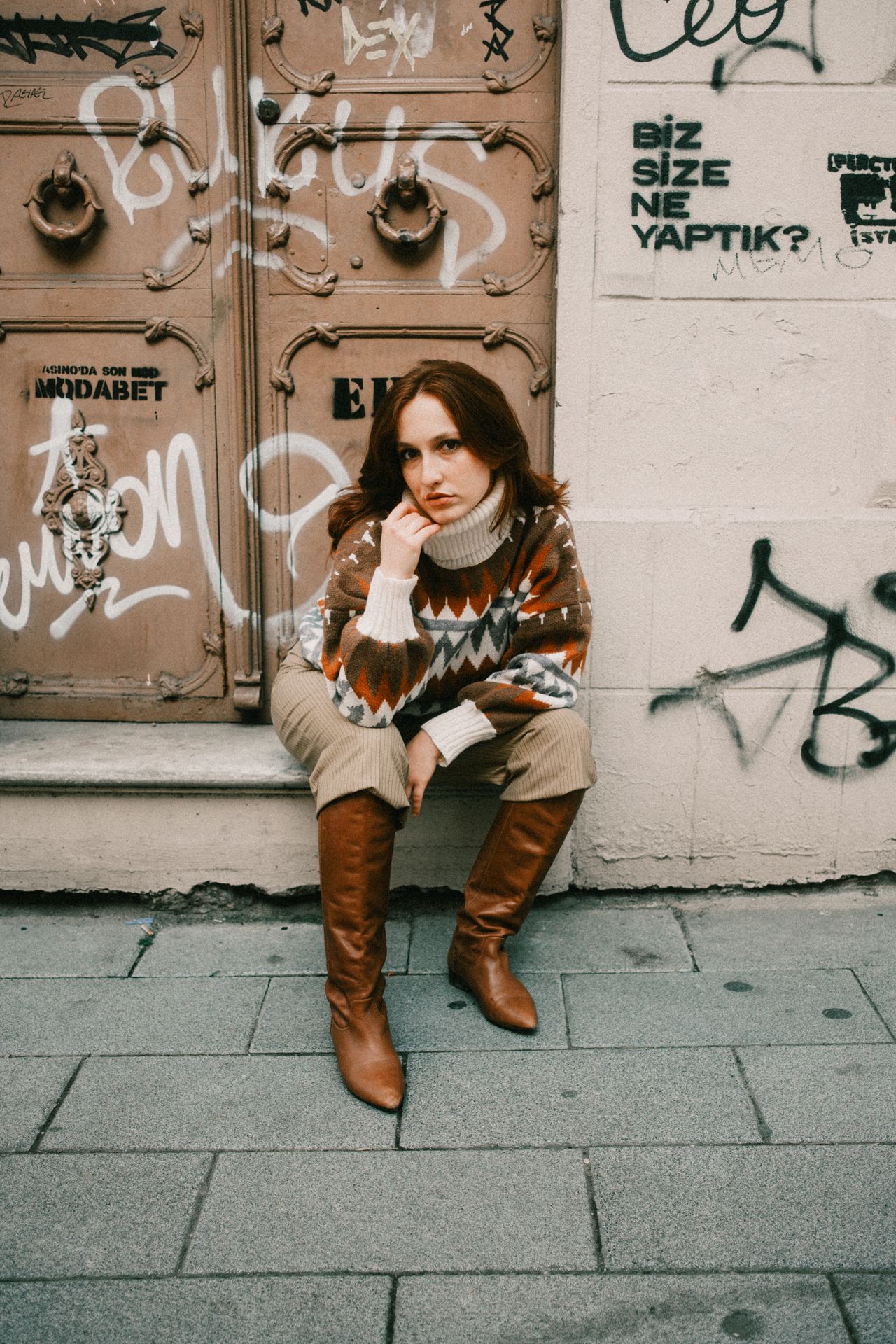 Woman in a brown and cream fair-isle knit sweater with tan knee boots sitting against a graffiti wall