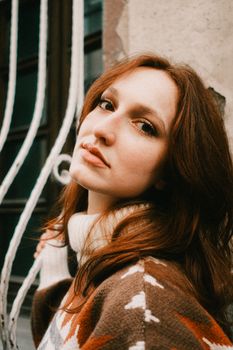 Fashionable woman with red hair posing in a cozy sweater outdoors by decorative railing.