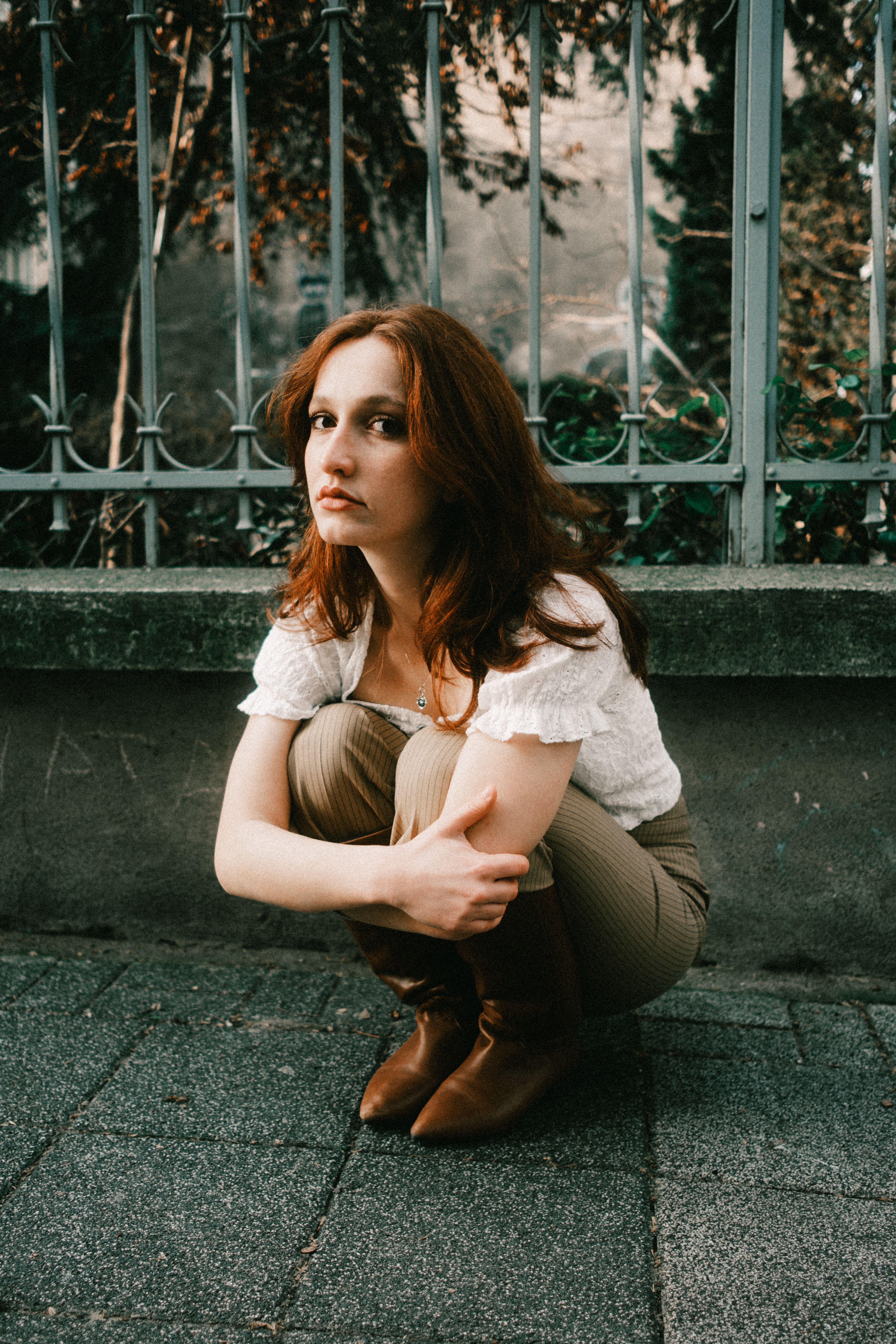 Young redheaded woman squatting by a fence, reflecting a thoughtful mood.
