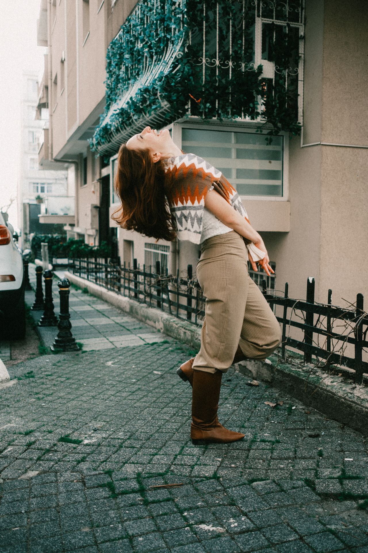 Woman in a patterned sweater vest and tan trousers posing playfully on a street